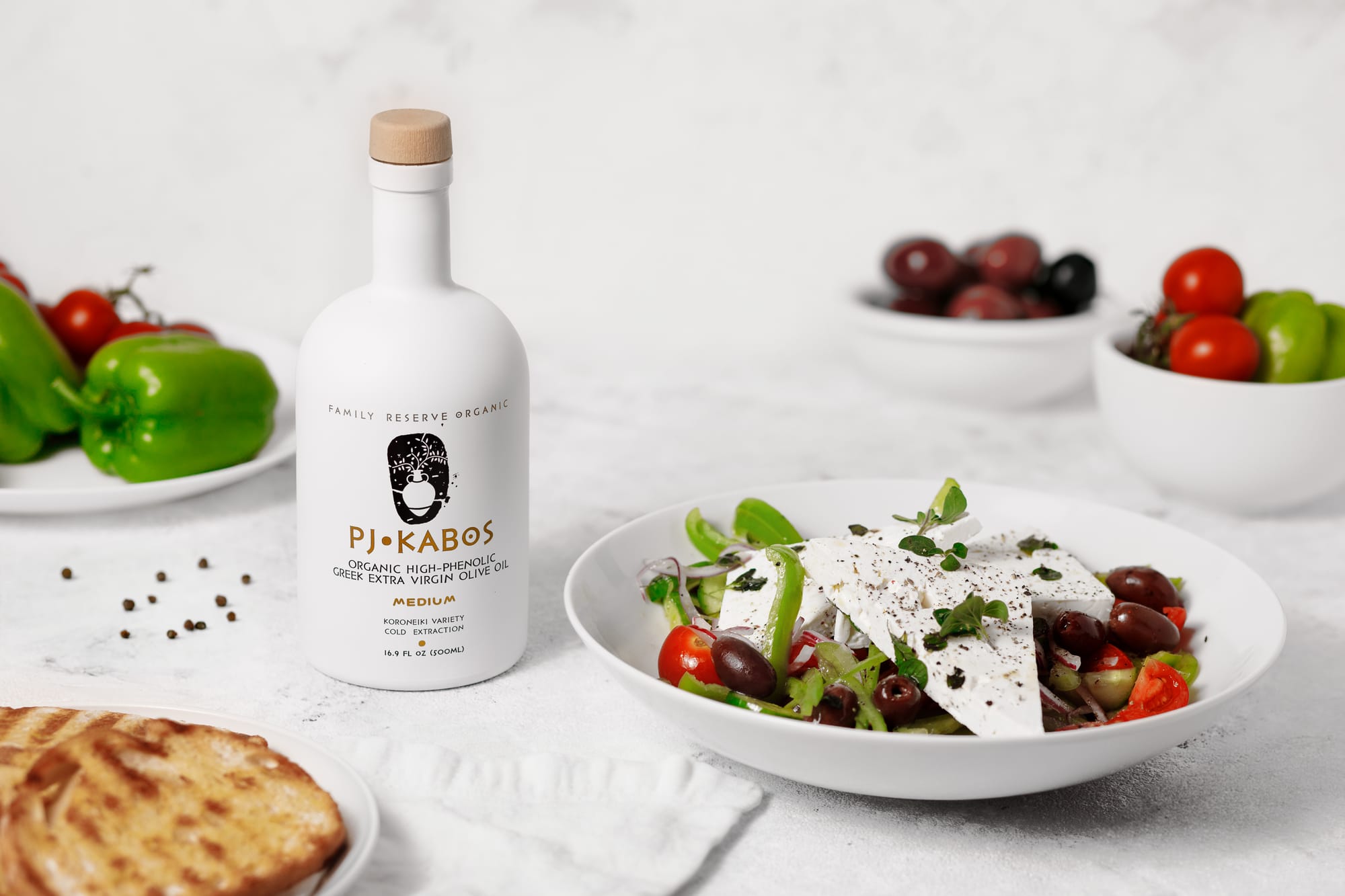 A bottle of high-phenolic extra virgin olive oil sitting on a table with a nutritious Greek Salad. Pita bread, peppers and olives are also on the table. Feta cheese is on the salad that has been drizzled with EVOO.
