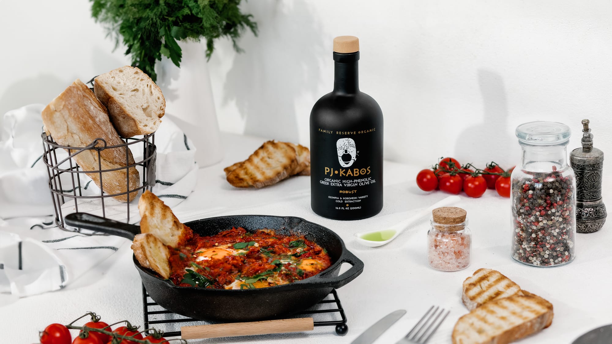 A Bottle of P Kabos Robust High-phenolic Extra Virgin Olive Oil on a table set for breakfast with the recently made tomatoes and eggs still in the skillet, fresh bread salt and pepper while a phenolic shot of oil rests in a spoon ready to be had before or during the meal.